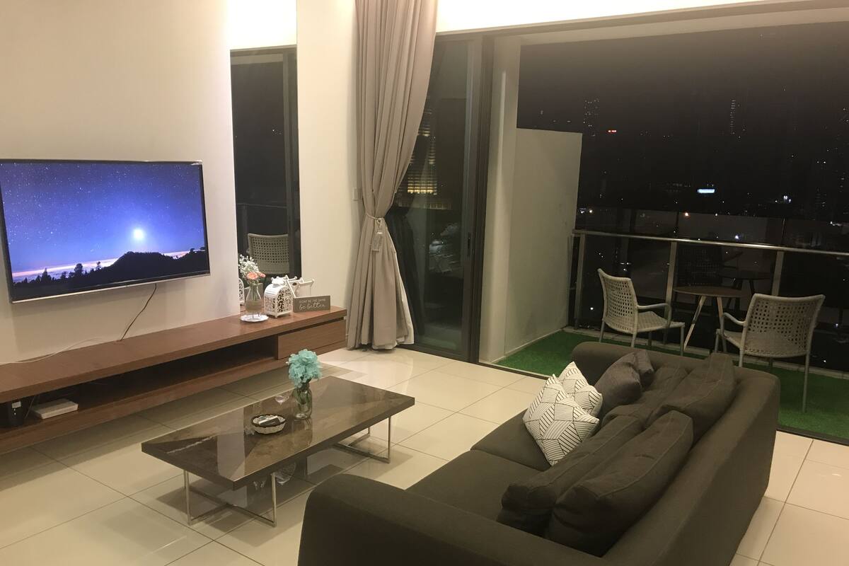 A modern living room with a large flat-screen TV, a glass coffee table, a dark-colored sofa, and a balcony with outdoor seating.