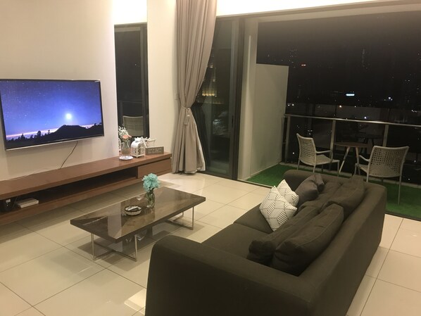 A modern living room with a large flat-screen TV, a glass coffee table, a dark-colored sofa, and a balcony with outdoor seating. - Setia Sky by Urban Homes (Kuala Lumpur)