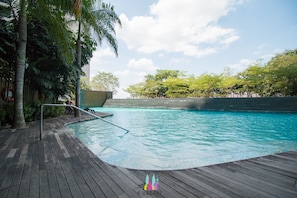 A wooden deck surrounding a swimming pool with palm trees and a view of a building in the background. - Setia Sky by Urban Homes (Kuala Lumpur)