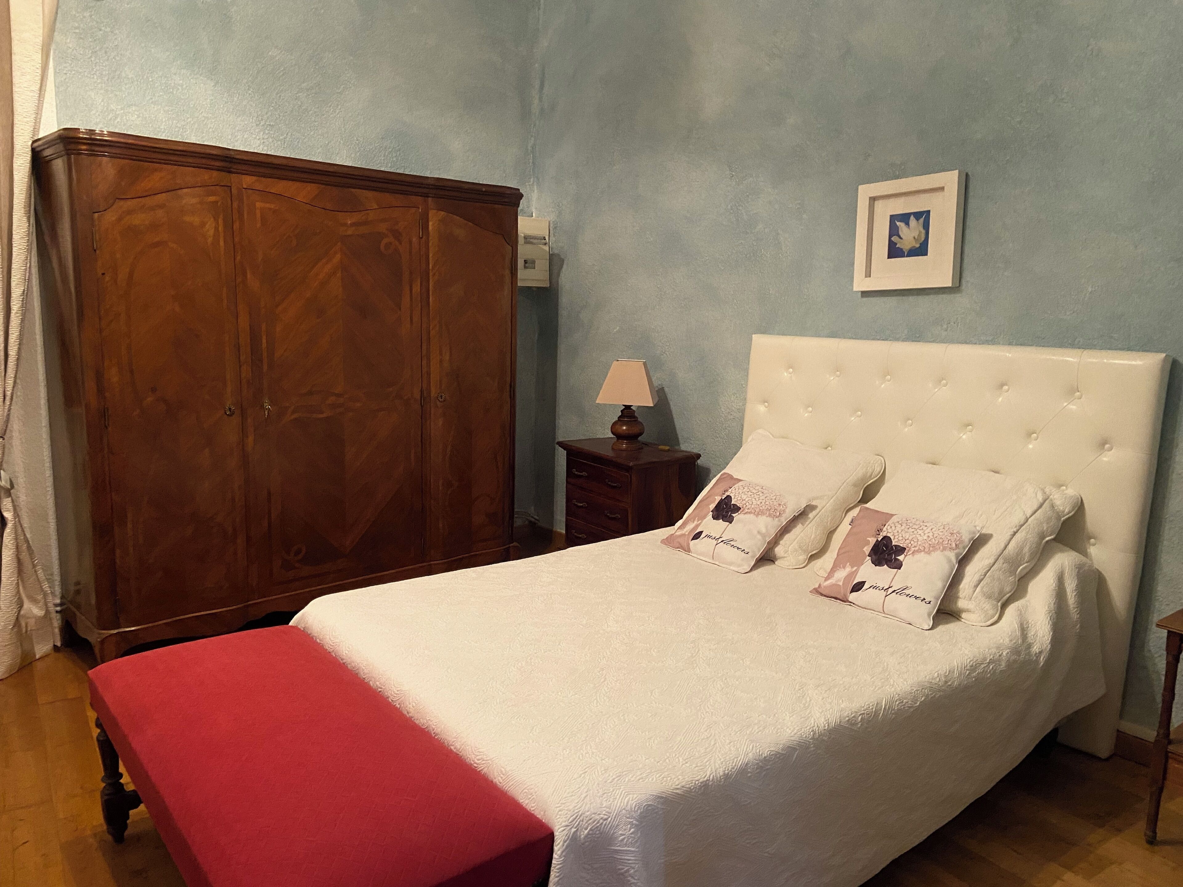 Een slaapkamer met een bed, een houten kledingkast, een nachtkastje met een lamp en een lijst met een afbeelding aan de muur.