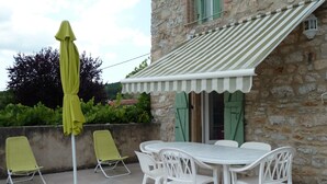 Une terrasse extérieure avec un mur en pierre, une table avec des chaises, un parasol jaune et un auvent à rayures.