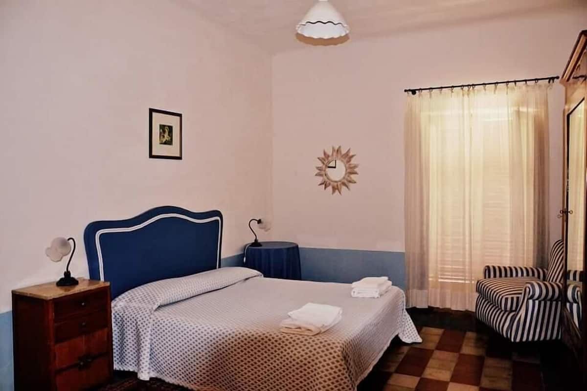 Een slaapkamer met een bed, nachtkastje, stoel, een wandklok en een raam met gordijnen.