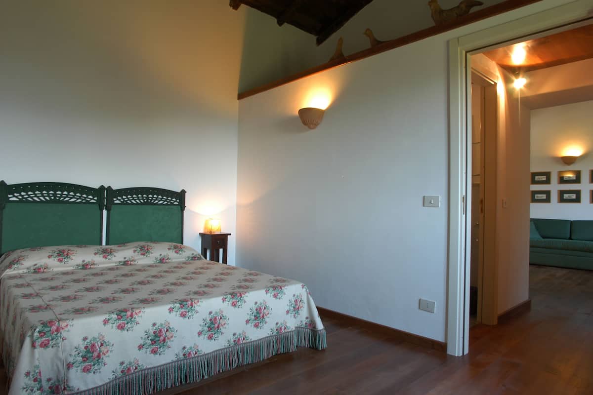 Een slaapkamer met een bloemenbedekking, een houten hoofdbord, een nachtkastje met een lamp, en een groene bank in de aangrenzende kamer.