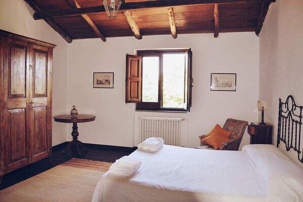 A bedroom with a wooden ceiling, a bed with white linens, a chair with an orange cushion, a small round table, and a window with an open shutter.