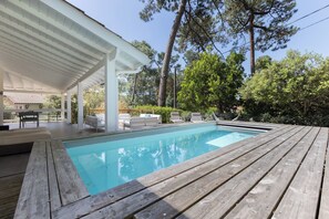 A swimming pool with a wooden deck, lounge chairs, and a covered patio area.