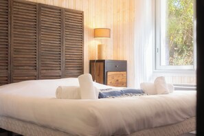 A bedroom with a large bed, wooden shutters, a nightstand, a lamp, and a window with a view of trees.