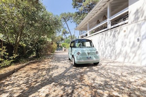 A vintage car parked on a cobblestone street, surrounded by trees and a white building with a balcony.