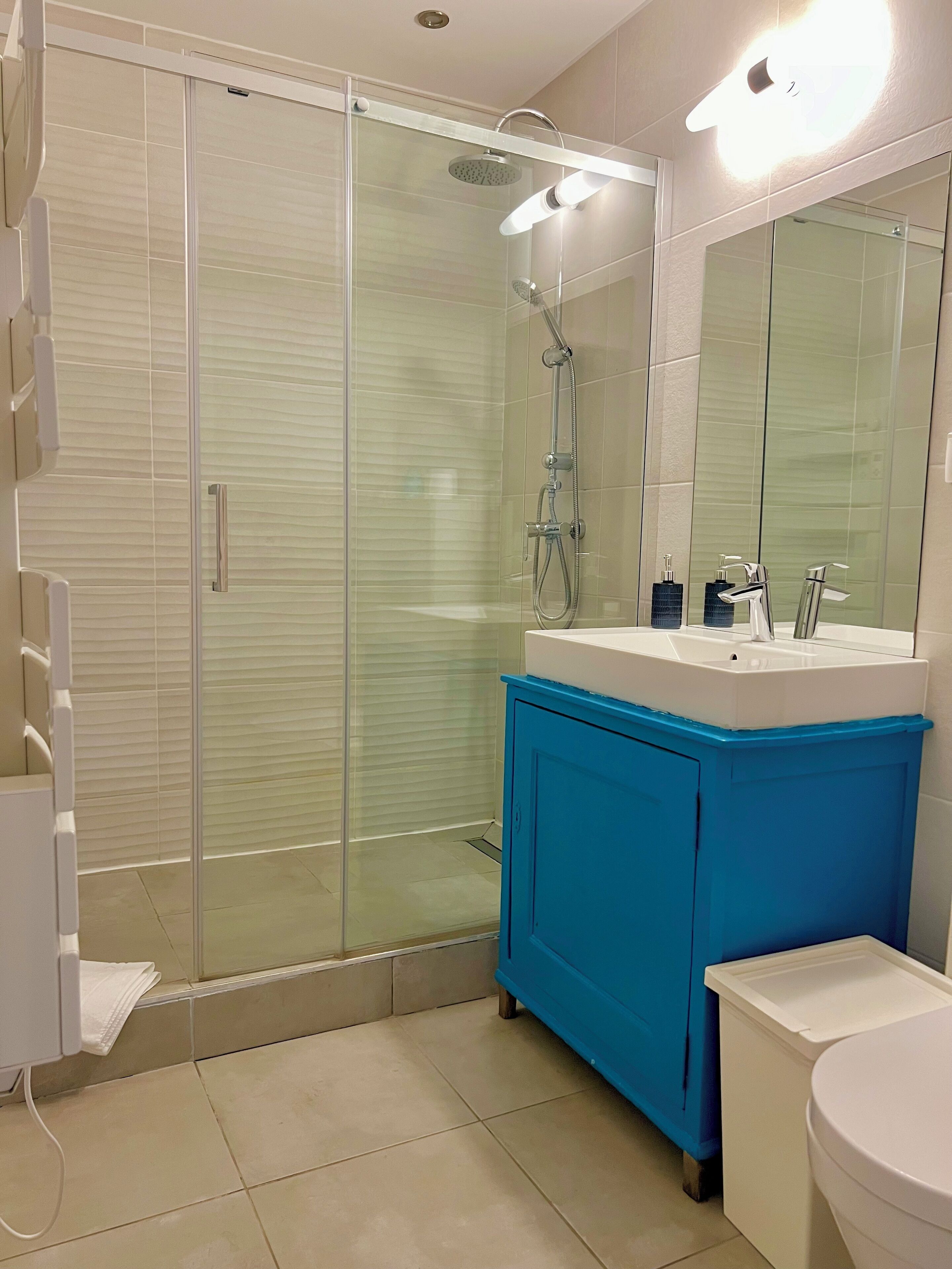 Un baño moderno con ducha de vidrio, un lavamanos blanco con gabinete azul y un espejo grande sobre el lavamanos.