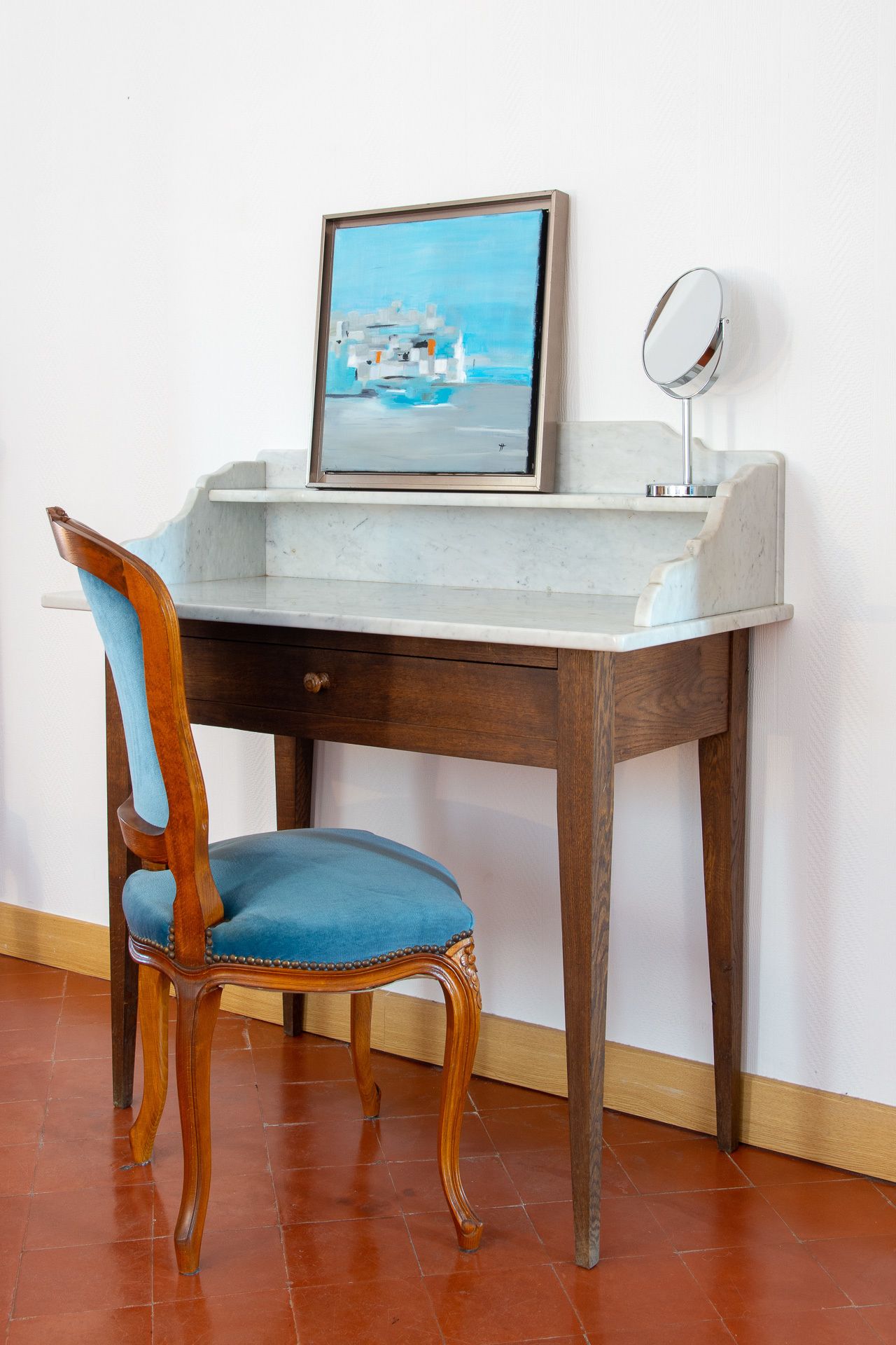 A wooden desk with a blue cushioned chair, a framed painting, and a mirror.