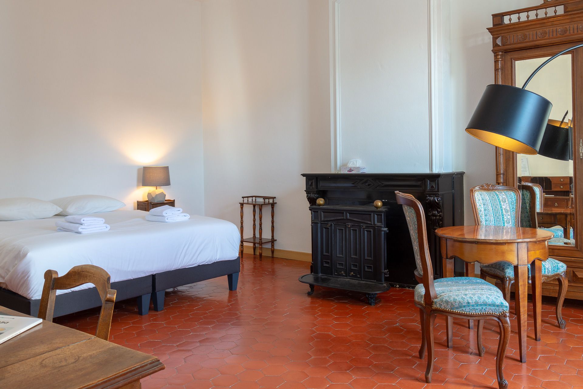 Quarto de hotel com cama, uma cadeira de madeira, uma mesinha, lareira e um espelho.