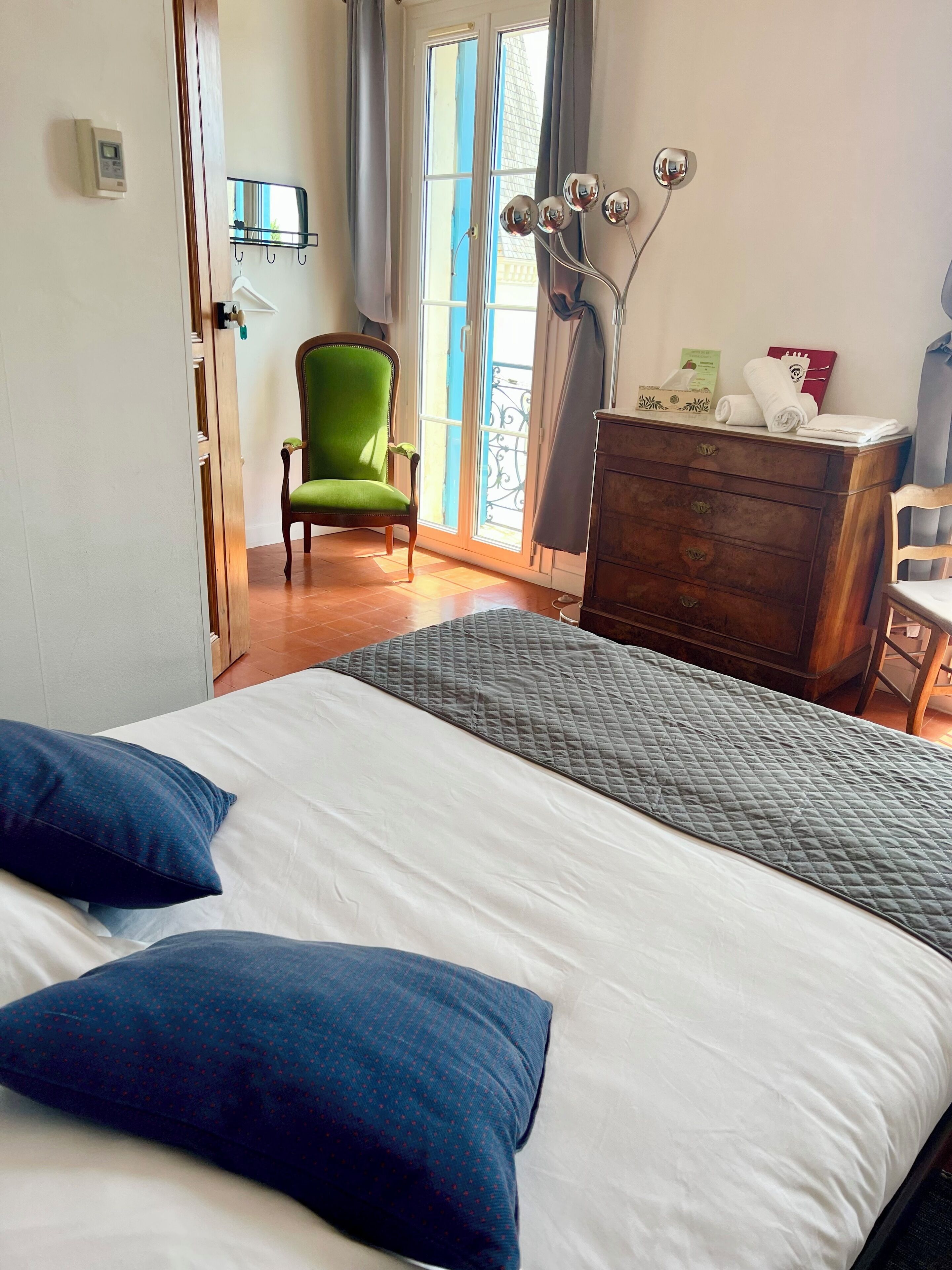 Une chambre d’hôtel avec un lit, un fauteuil vert, une commode en bois et une fenêtre avec des rideaux.