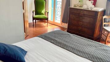Une chambre d’hôtel avec un lit, un fauteuil vert, une commode en bois et une fenêtre avec des rideaux.