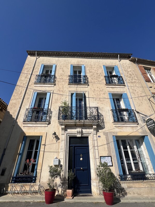 A multi-story building with a central entrance, balconies with wrought iron railings, and potted plants at the entrance. - Entre MIDI - Maison d'Hôtes - B&B (Capestang)