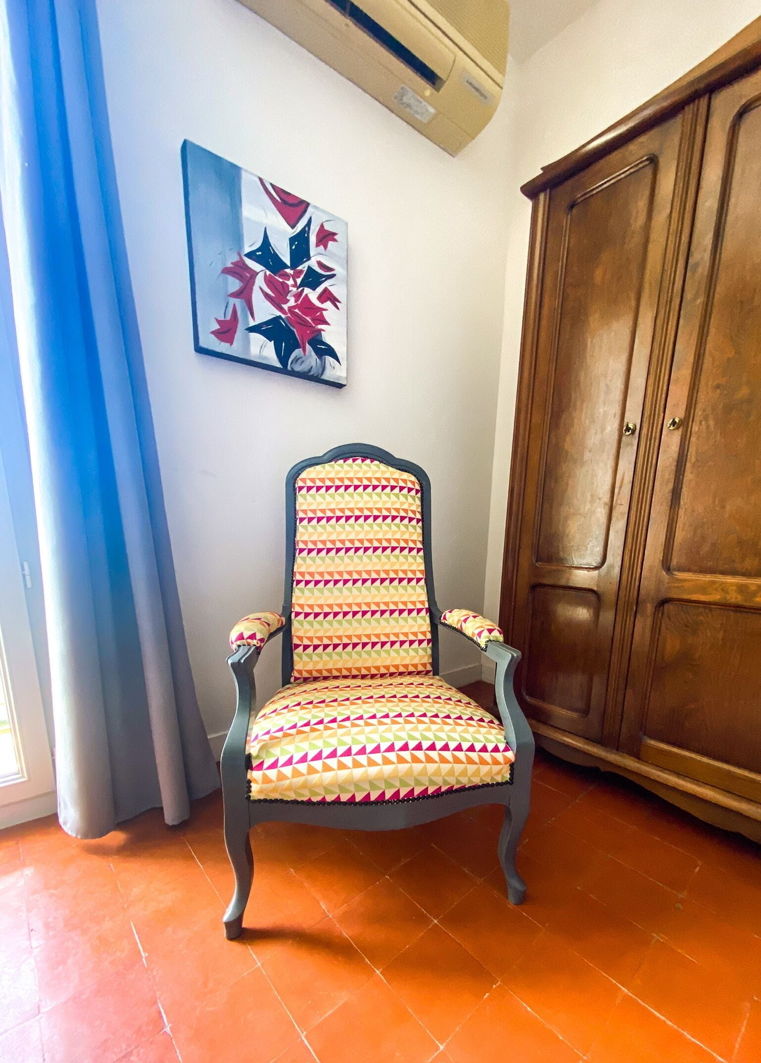A chair with a geometric patterned cushion, a wooden wardrobe, a wall-mounted air conditioner, and a blue curtain.