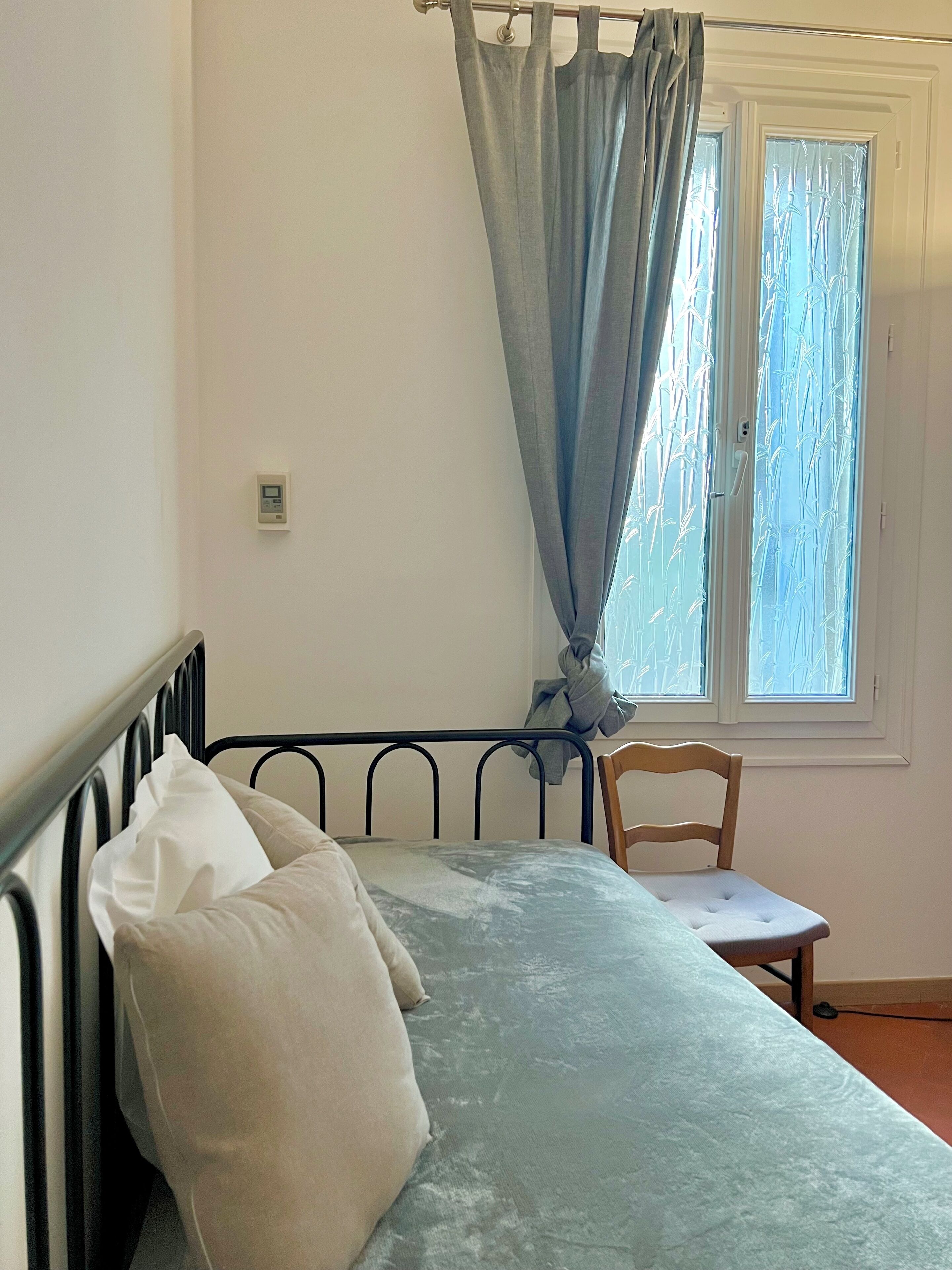 Ein Schlafzimmer mit Bett, Sessel, einem Fenster mit Milchglas und einem Thermostat an der Wand.