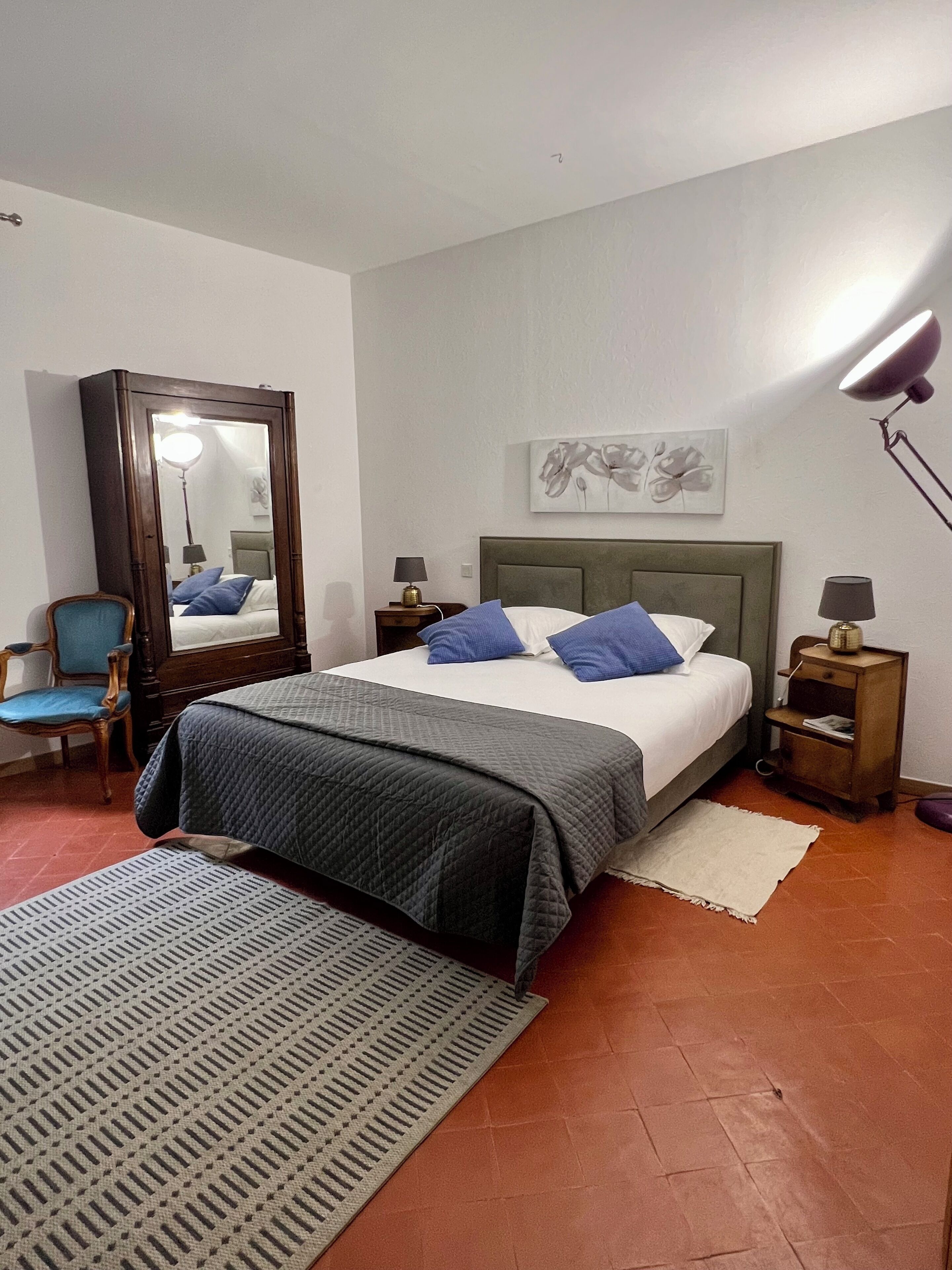 Un dormitorio con cama, una silla, un espejo, una lámpara y una obra de arte en la pared.
