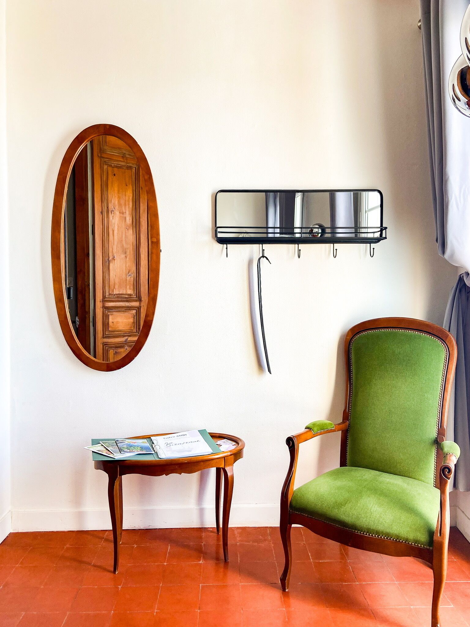 A room with a wooden door, a green upholstered chair, a small wooden table with magazines, a wall-mounted mirror, and a black metal shelf.