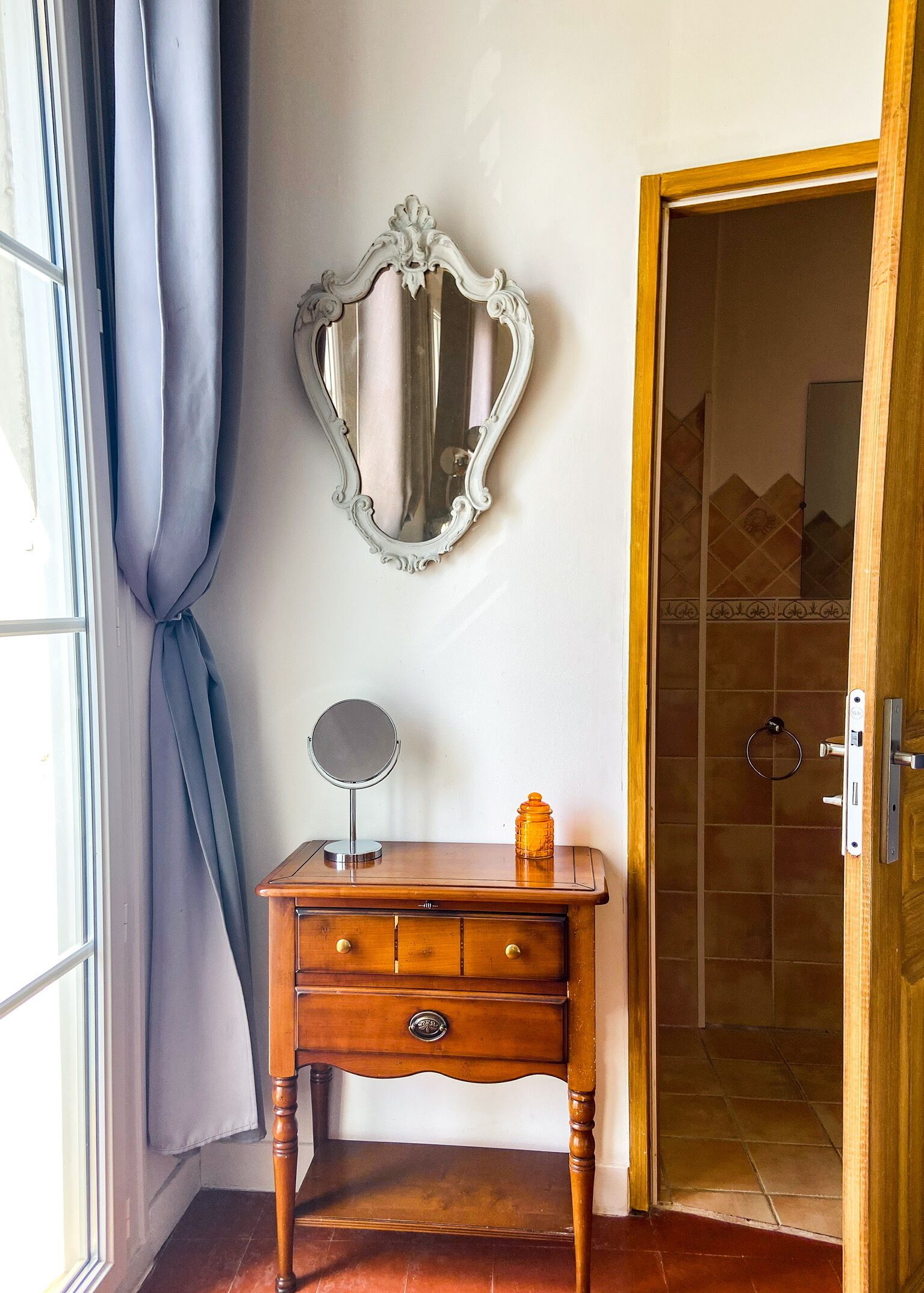 A wooden dresser with a mirror and a small bottle, a decorative mirror on the wall, and a door leading to a tiled bathroom.