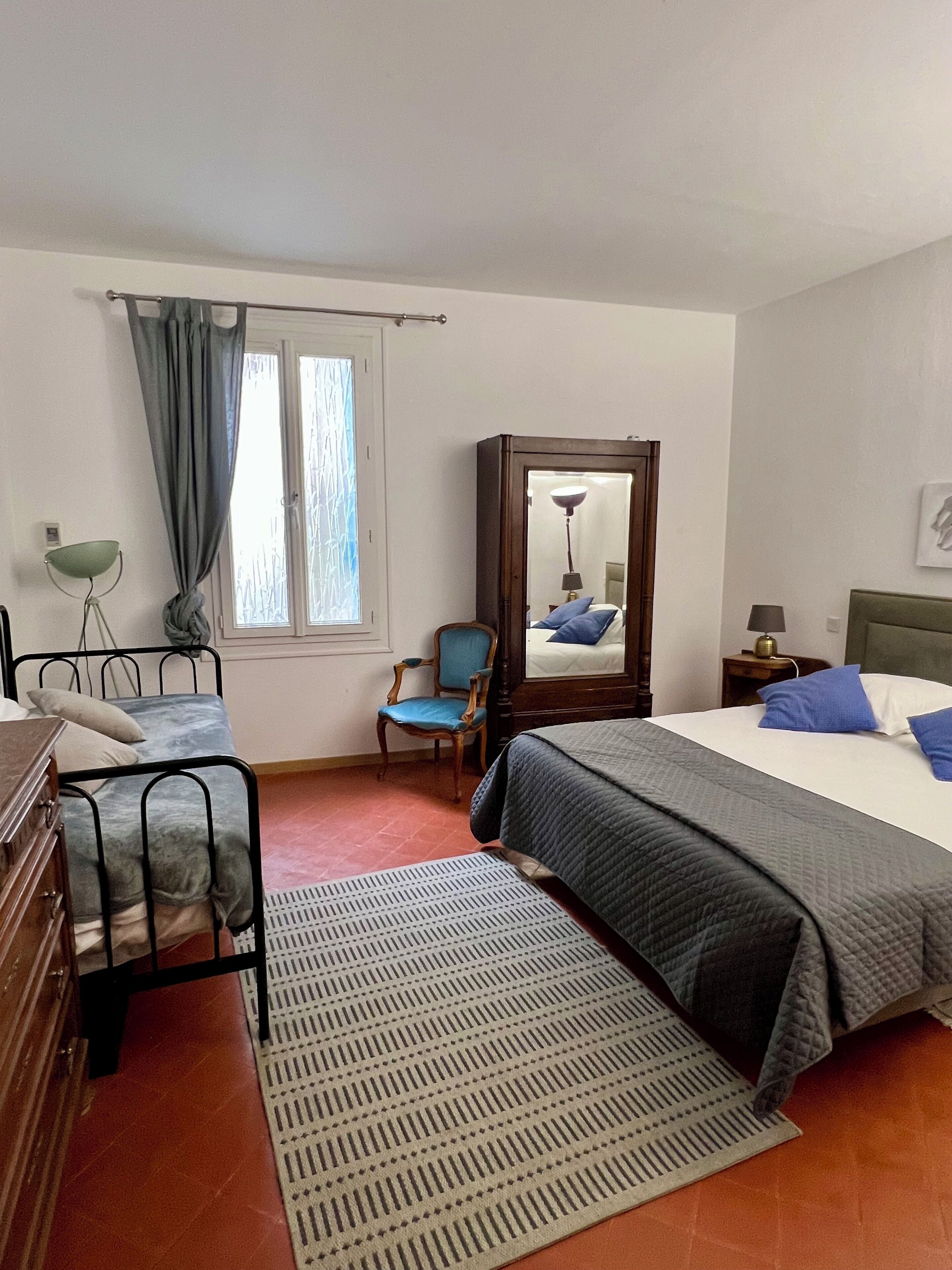 Une chambre à coucher avec un lit, une chaise, un bureau, un miroir et une fenêtre avec des rideaux.