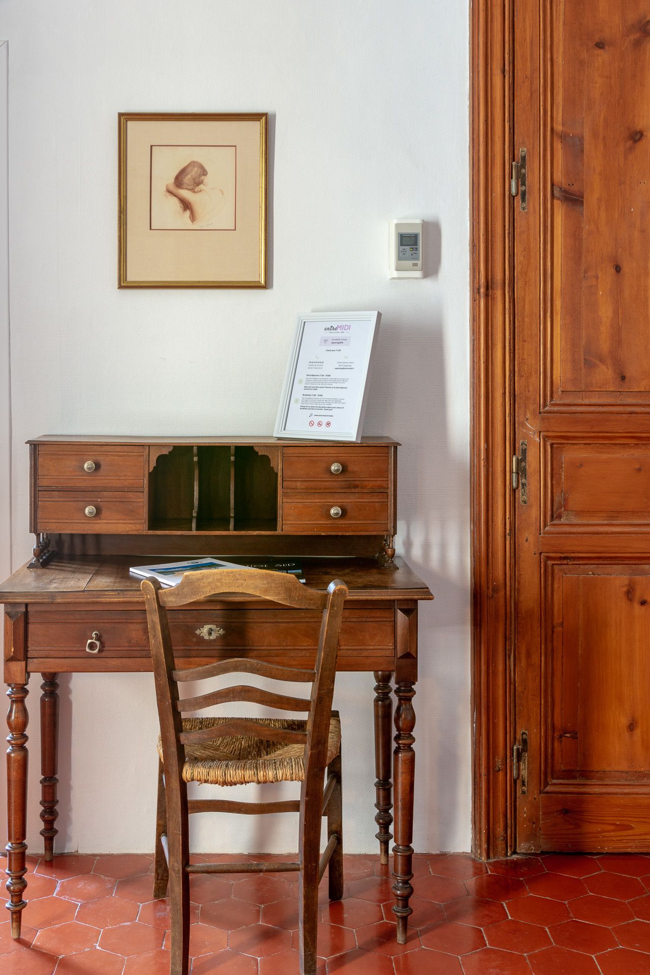 A wooden desk with drawers, a chair with a woven seat, a framed picture on the wall, and a wooden cabinet.