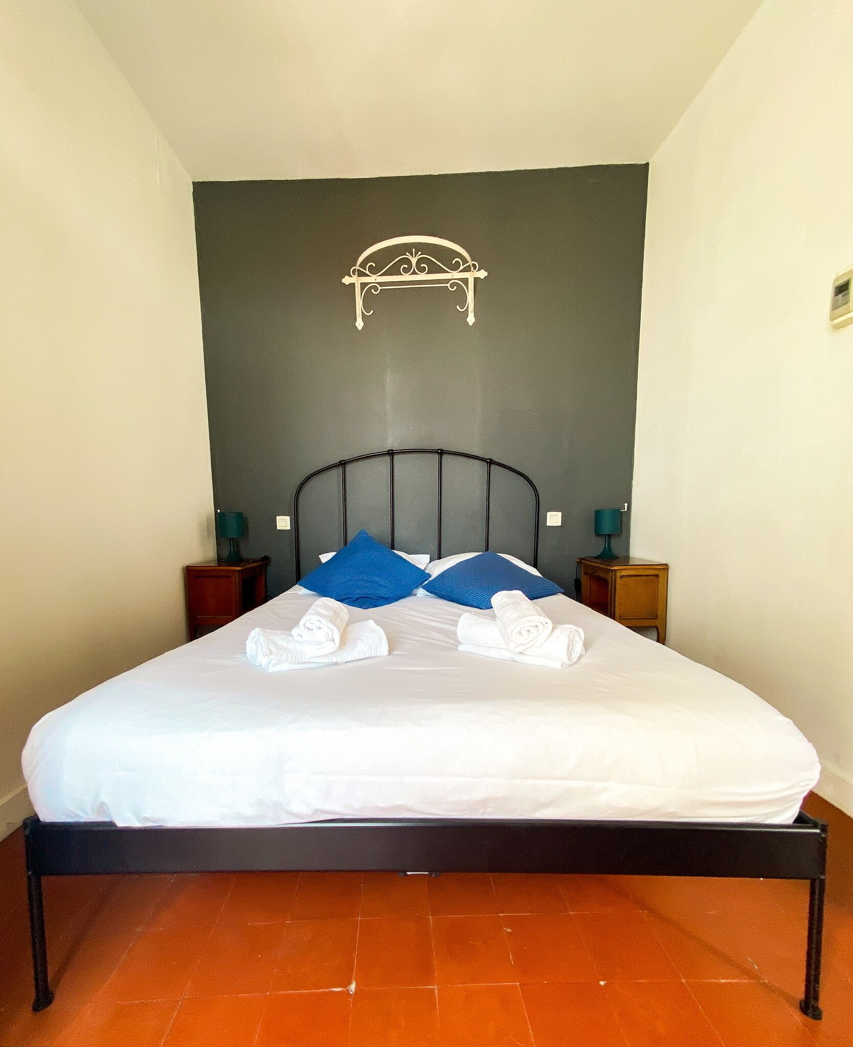 Un lit bien fait, avec du linge de lit blanc et des oreillers bleus, une tête de lit en métal noir et deux tables de chevet avec des lampes.