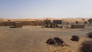 A desert landscape with several buildings, including a large structure with a flat roof and smaller buildings with flat roofs. There are palm trees and a pile of sticks in the foreground. - Riad Desert Camel (Taouz)