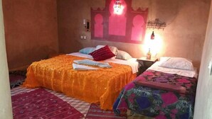 A room with two beds, one with an orange bedspread and the other with a floral bedspread. There is a patterned rug on the floor, a wall-mounted light fixture, and a small table with a bottle on it. - Riad Desert Camel (Taouz)