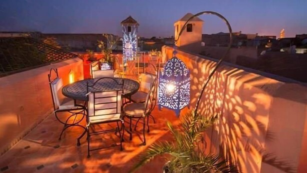 An outdoor patio with a round table and chairs, a decorative lantern, and a view of the city skyline at dusk. - Riad Desert Camel (Taouz)