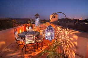 An outdoor patio with a round table and chairs, a decorative lantern, and a view of the city skyline at dusk. - Riad Desert Camel (Taouz)