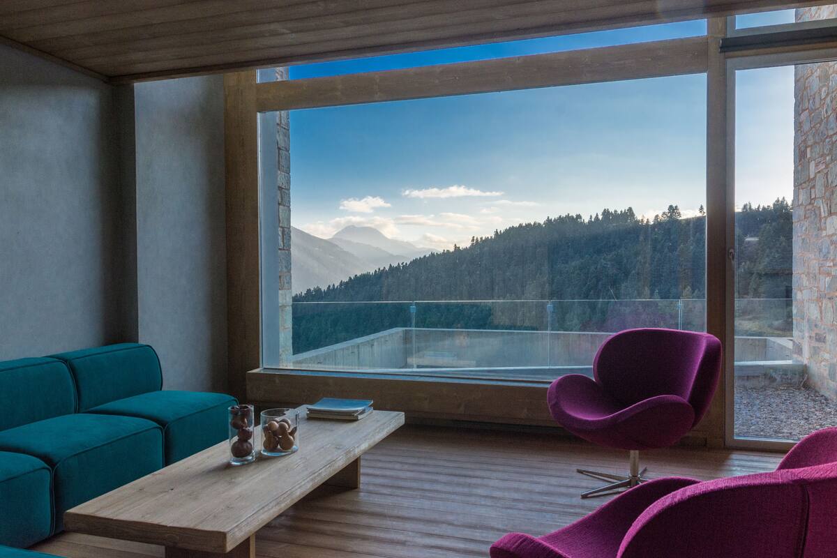 A modern room with a wooden floor, teal sofa, and a purple chair. A wooden table with a book and glass is in front of a large window overlooking a mountainous landscape.