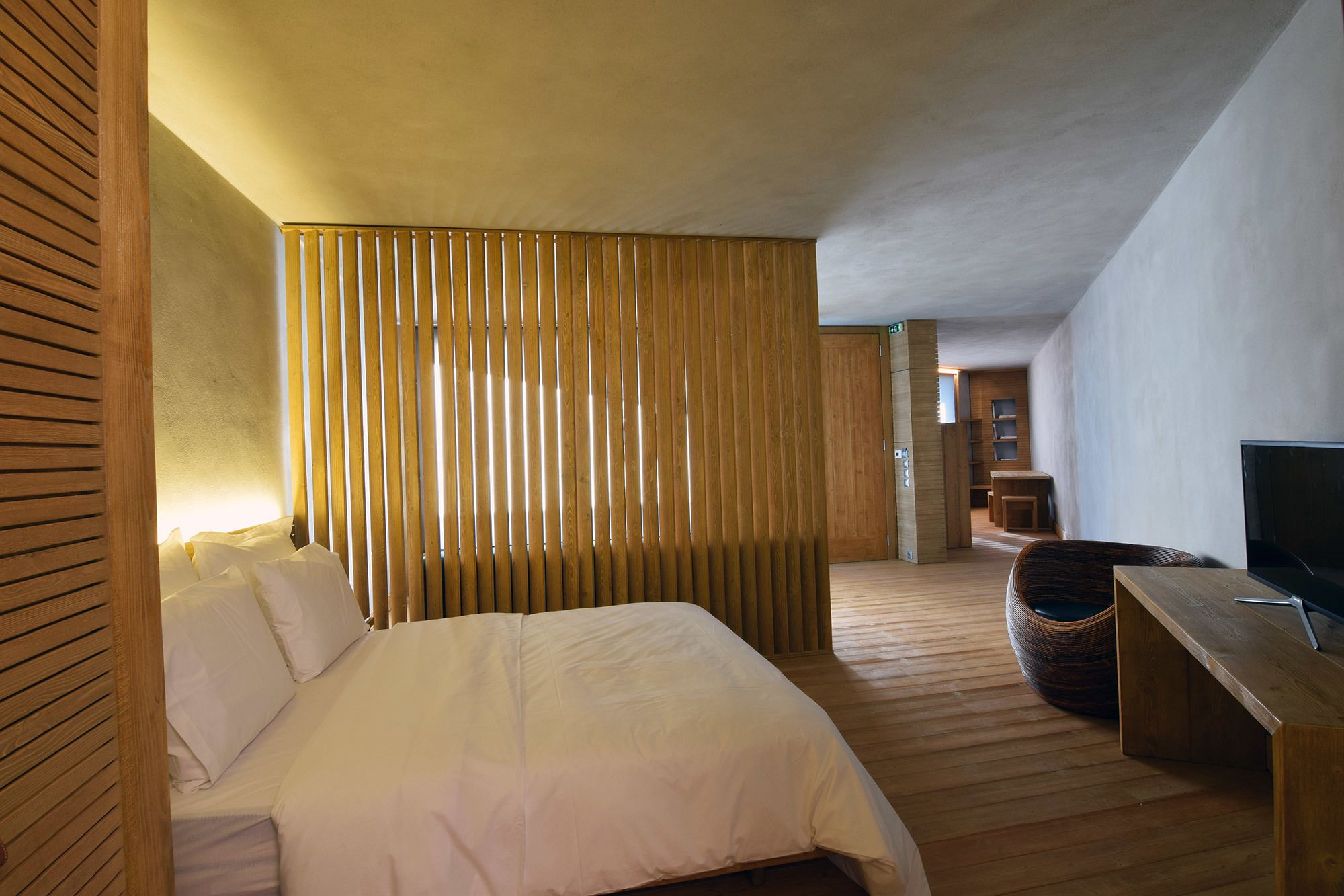 A hotel room with a bed, a wooden partition, a television, and a wicker chair.