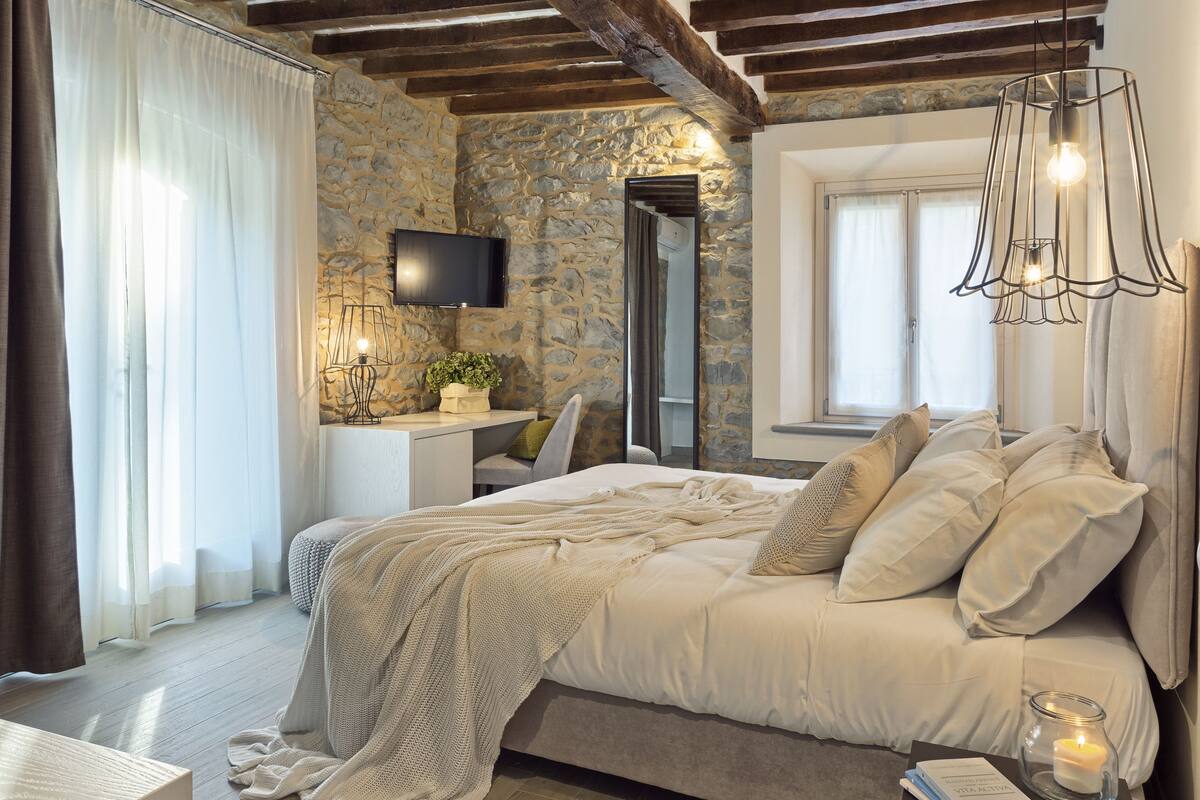 A bedroom with a stone wall, a bed with white bedding, a bedside table with a candle, and a TV mounted on the wall.
