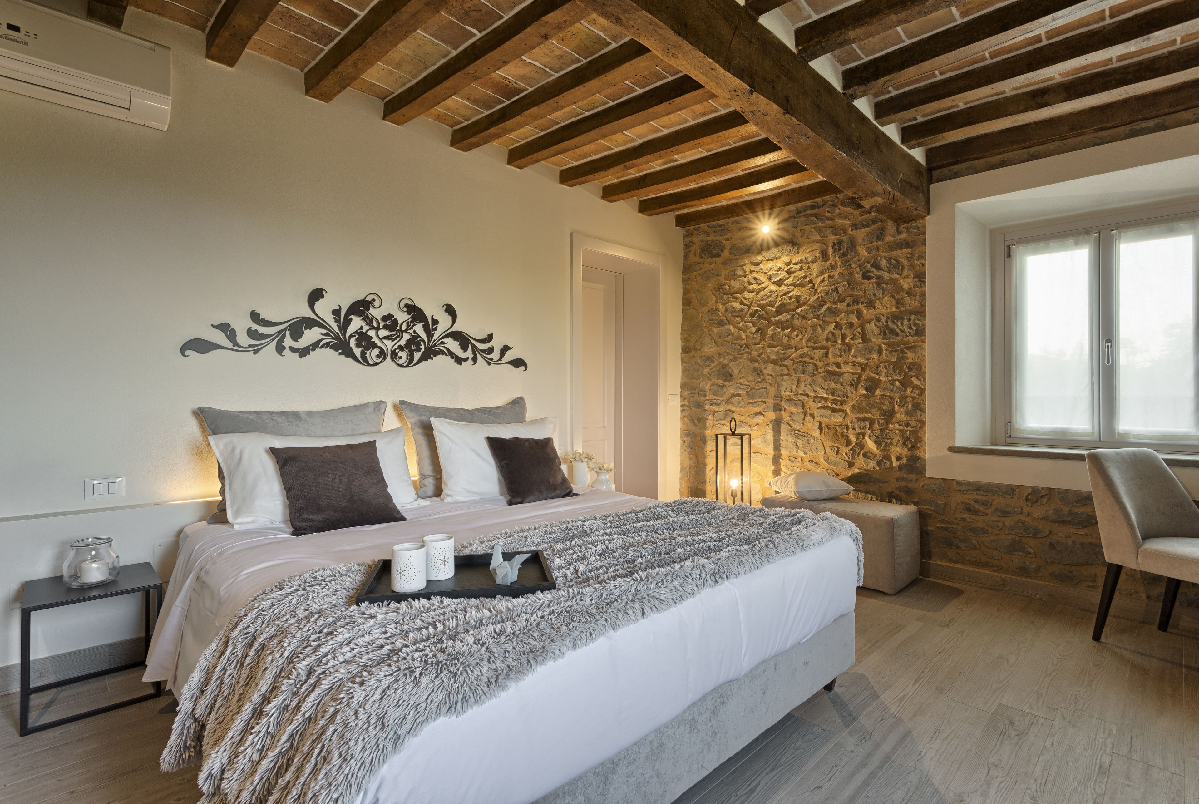 A bedroom with a stone wall, wooden beams, a bed with a fluffy blanket, bedside tables, and a chair.