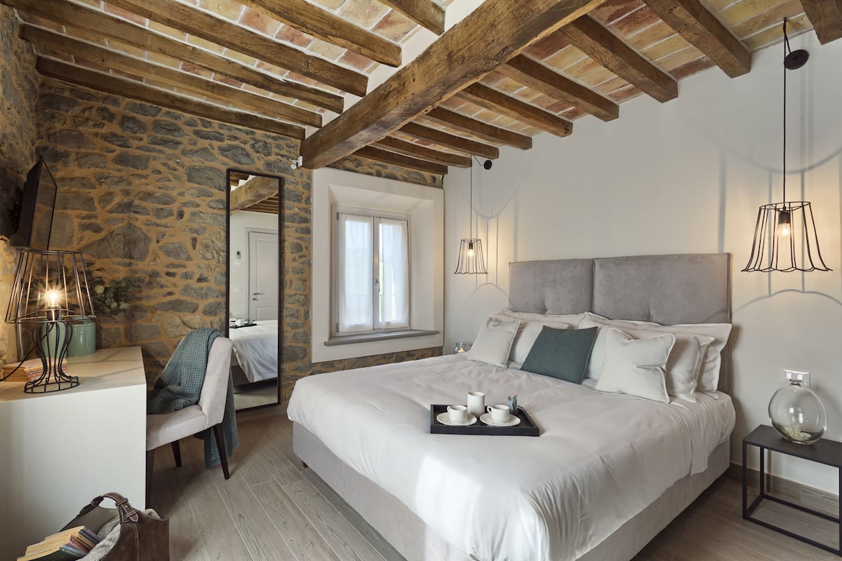 A bedroom with a stone wall, wooden beams, a bed with white bedding, a bedside table with a lamp, and a window with curtains.