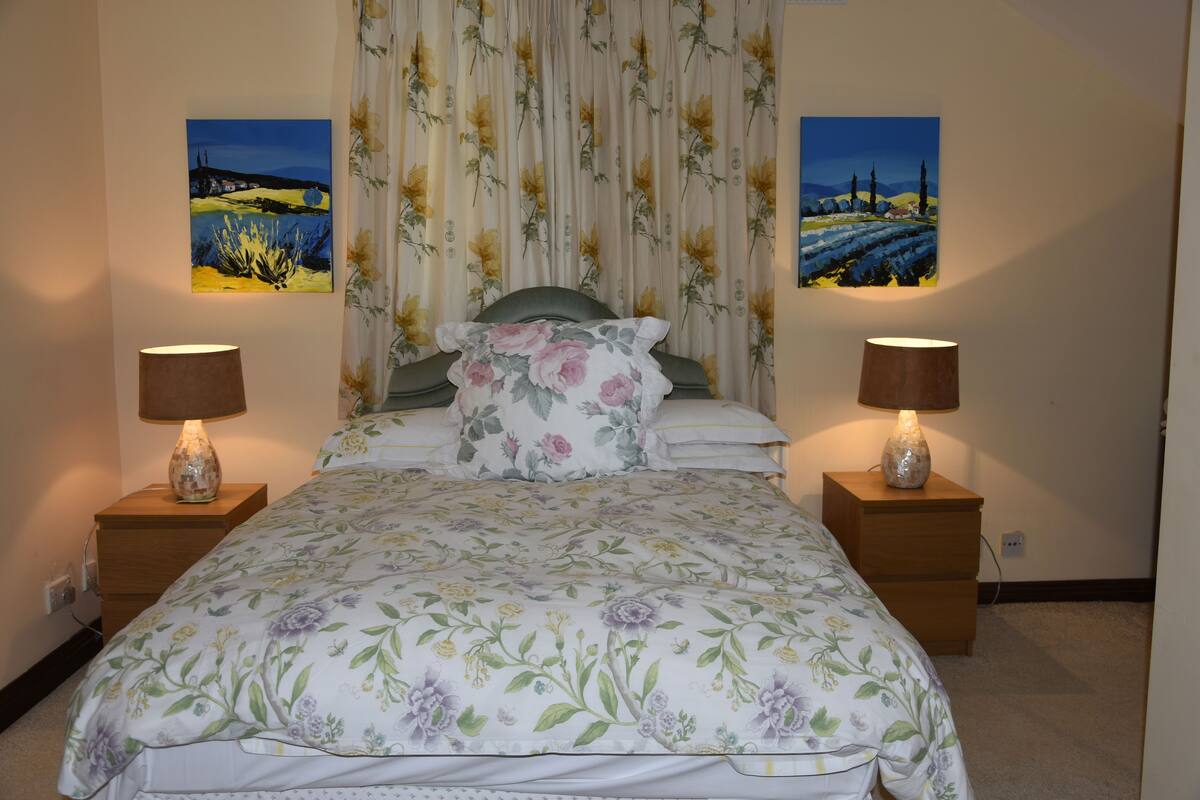 A bedroom with a floral bedspread, two bedside lamps, a headboard, and two landscape paintings on the walls.