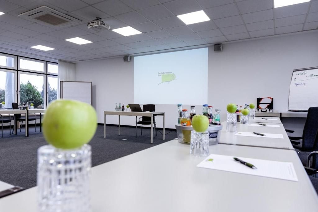 Un salón de conferencias con una pantalla de proyector, mesas con botellas de agua y una pizarra.