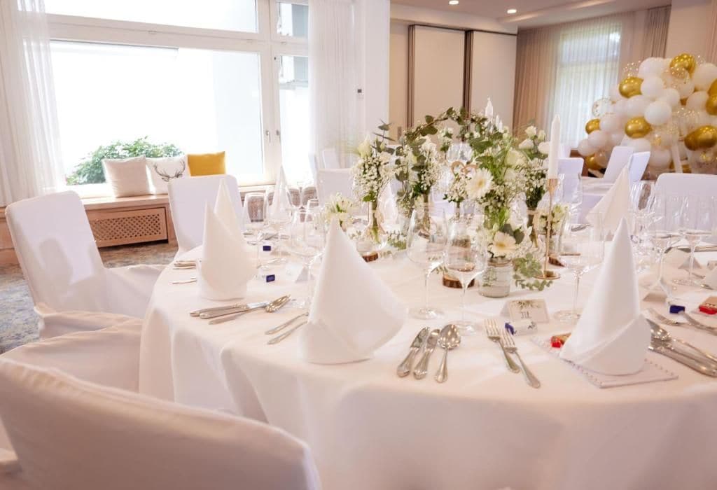 Una mesa de banquete dispuesta con manteles blancos, cubertería y un centro de mesa con flores.