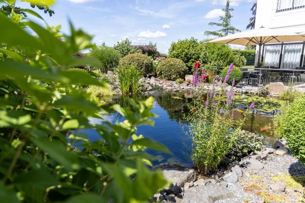 Un jardín tranquilo con un estanque, diversas plantas y un edificio con mesas al aire libre.