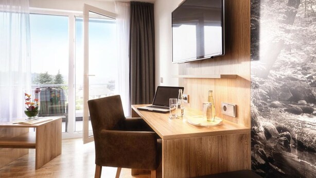 A modern hotel room with a wooden desk, a laptop, a flat-screen TV mounted on the wall, and a large window offering a view of the outdoors. - Hotel Derichsweiler Hof (Nümbrecht)