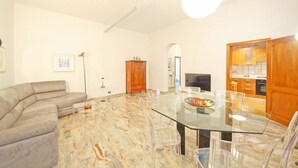 A modern living room with a curved gray sofa, a glass dining table with clear chairs, a wooden cabinet, and a kitchen area visible through an archway. - DolceVita Apartments N. 250 (Venice)
