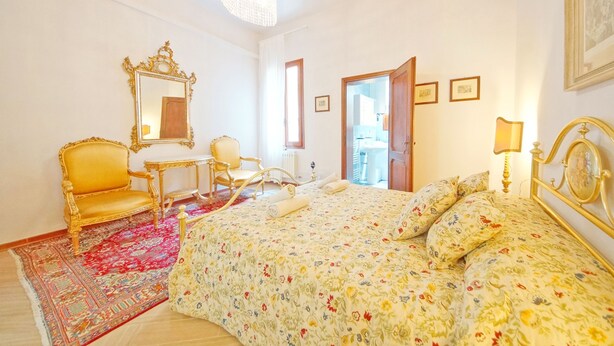 A hotel room with a bed, a golden chair, a small table, a mirror, and a door leading to another room. - DolceVita Apartments N. 250 (Venice)