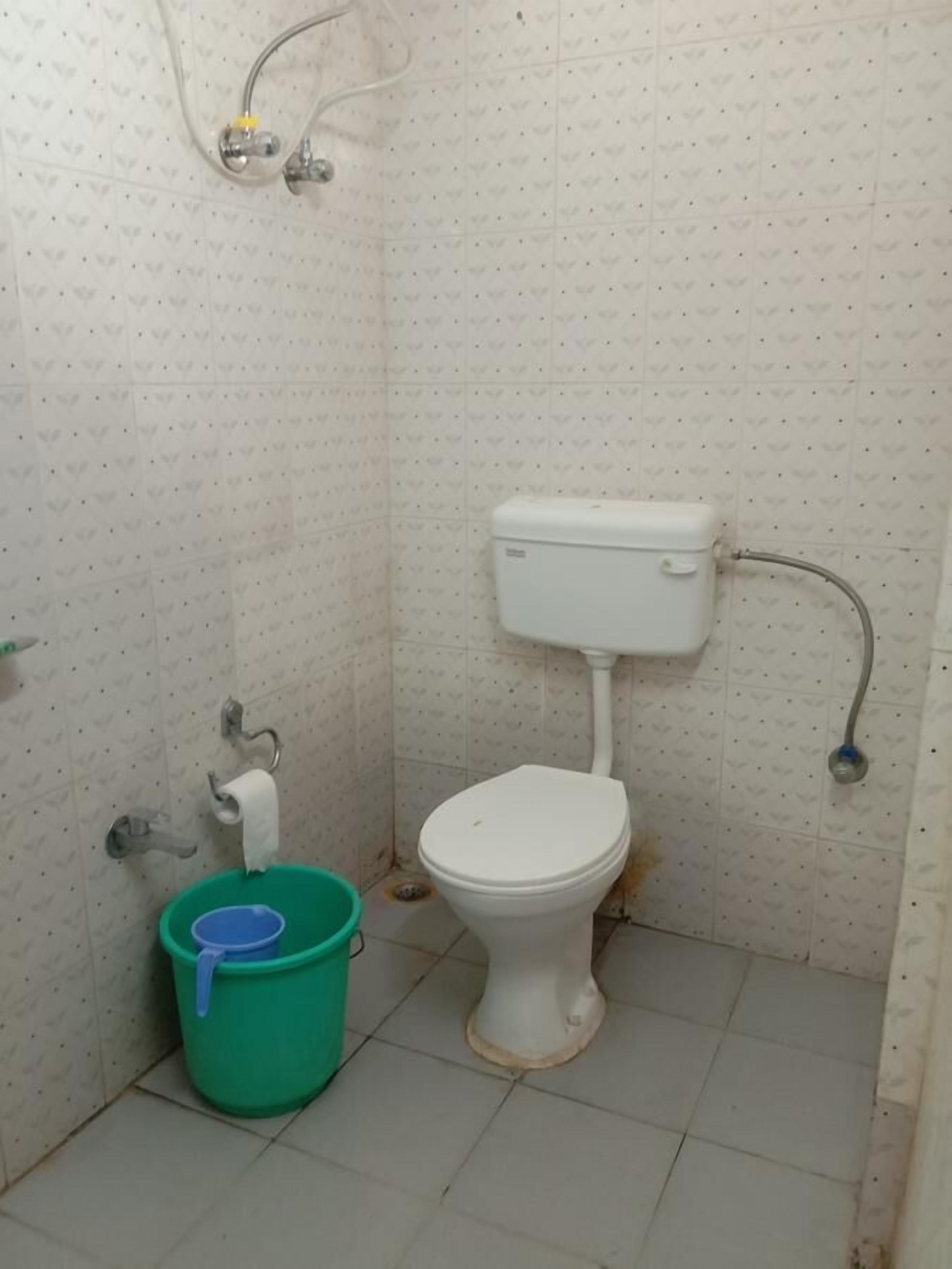 Une petite salle de bain simple, avec des toilettes, un seau vert et un tuyau de douche.