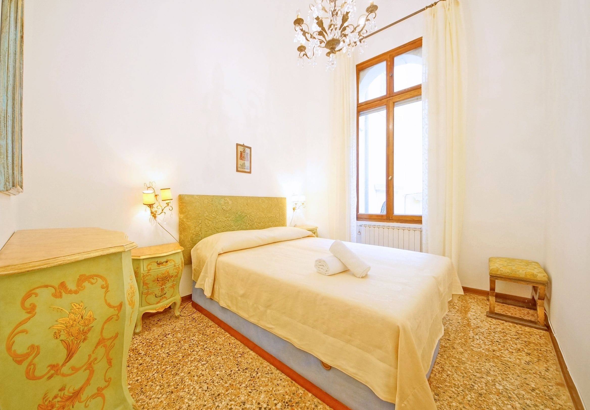 A bedroom with a bed, bedside lamps, a small table, a bench, and a window with curtains.