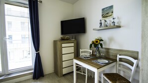 A room with a wooden desk, white chairs, a television, a vase with flowers, and a window with curtains. - Baltic-Apartments - Aquamarina C12 (Swinoujscie)