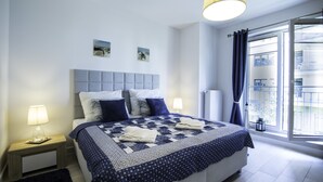 A modern bedroom with a large bed, a headboard, bedside lamps, and a view of the outside through a window. - Baltic-Apartments - Aquamarina C12 (Swinoujscie)
