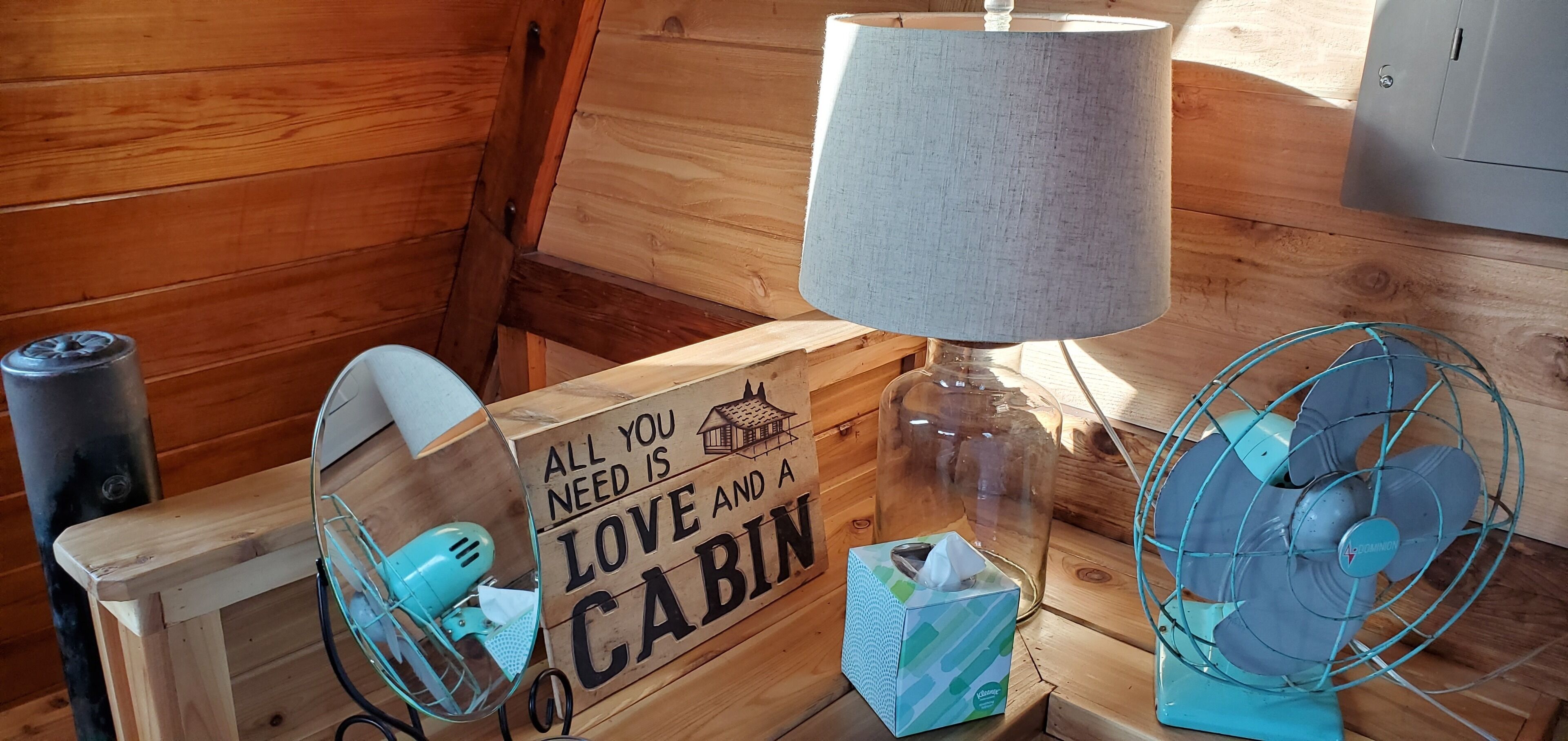 A wooden cabin interior with a wooden table, a lamp, a fan, a tissue box, and a sign that reads "ALL YOU NEED IS LOVE AND A CABIN."