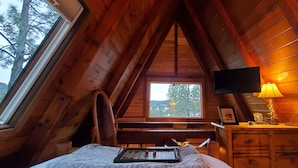 Une chambre confortable située dans les combles, avec un plafond en pente en bois, un lit recouvert d’une couette blanche, une table de chevet avec une lampe et un téléviseur fixé au mur.