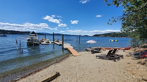Une jetée au bord du lac avec des bateaux, une plateforme en bois et une plage de galets agrémentée de chaises longues et d’un parasol.