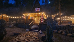 Un chalet doté d’un escalier en pierre menant à une terrasse en bois, entouré de guirlandes lumineuses et offrant une vue sur un paysage montagneux.