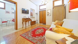 Ein Wohnzimmer mit weißem Sofa, einem Teppich mit roten und goldenen Mustern, Holzmöbeln, einem an der Wand befestigten Fernseher und einer Kristallleuchtre.